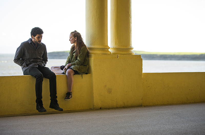 TV series Oridinary Lies 2 - Toke (Joel Fry) & Chrissy (Ella Peel) sat on yellow wall by sea talking